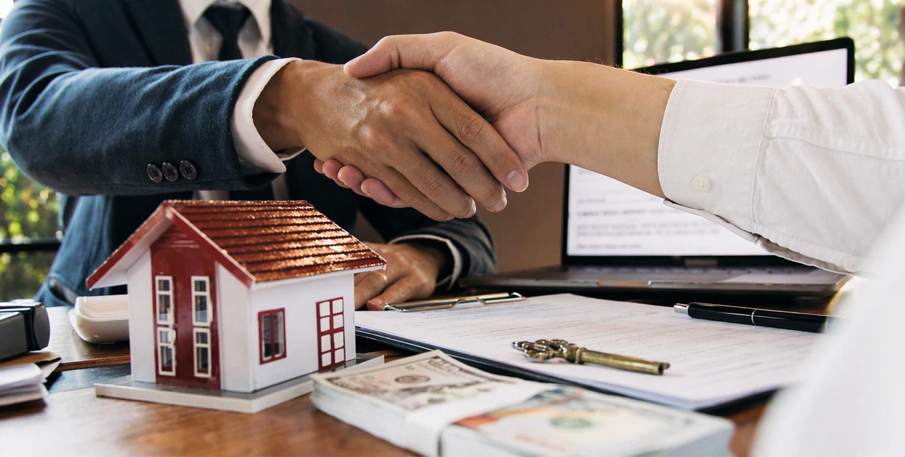 Two people shaking hands over a signed agreement, with a model house and keys on the desk